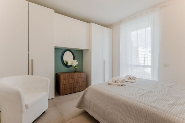A bedroom with a bed, a white armchair, a wooden dresser, and a mirror.