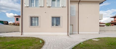 A two-story building with a paved walkway, two windows with shutters, and a small balcony.