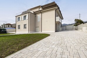 A modern two-story house with a paved driveway, a small lawn, and a mountain backdrop. - GuestHost - Mare Monti Apartment 5 (Dogana)