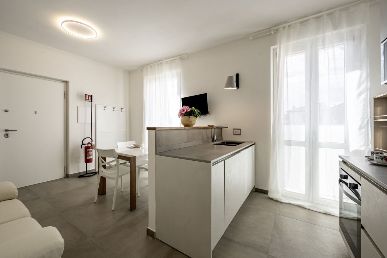 A modern kitchen with a central island, a dining area with a white table and chairs, a sofa, and a TV mounted on the wall.