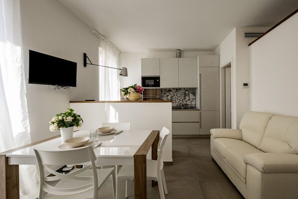 A modern kitchen with white cabinets, a microwave, and a refrigerator. There is a dining table with chairs and a TV mounted on the wall.