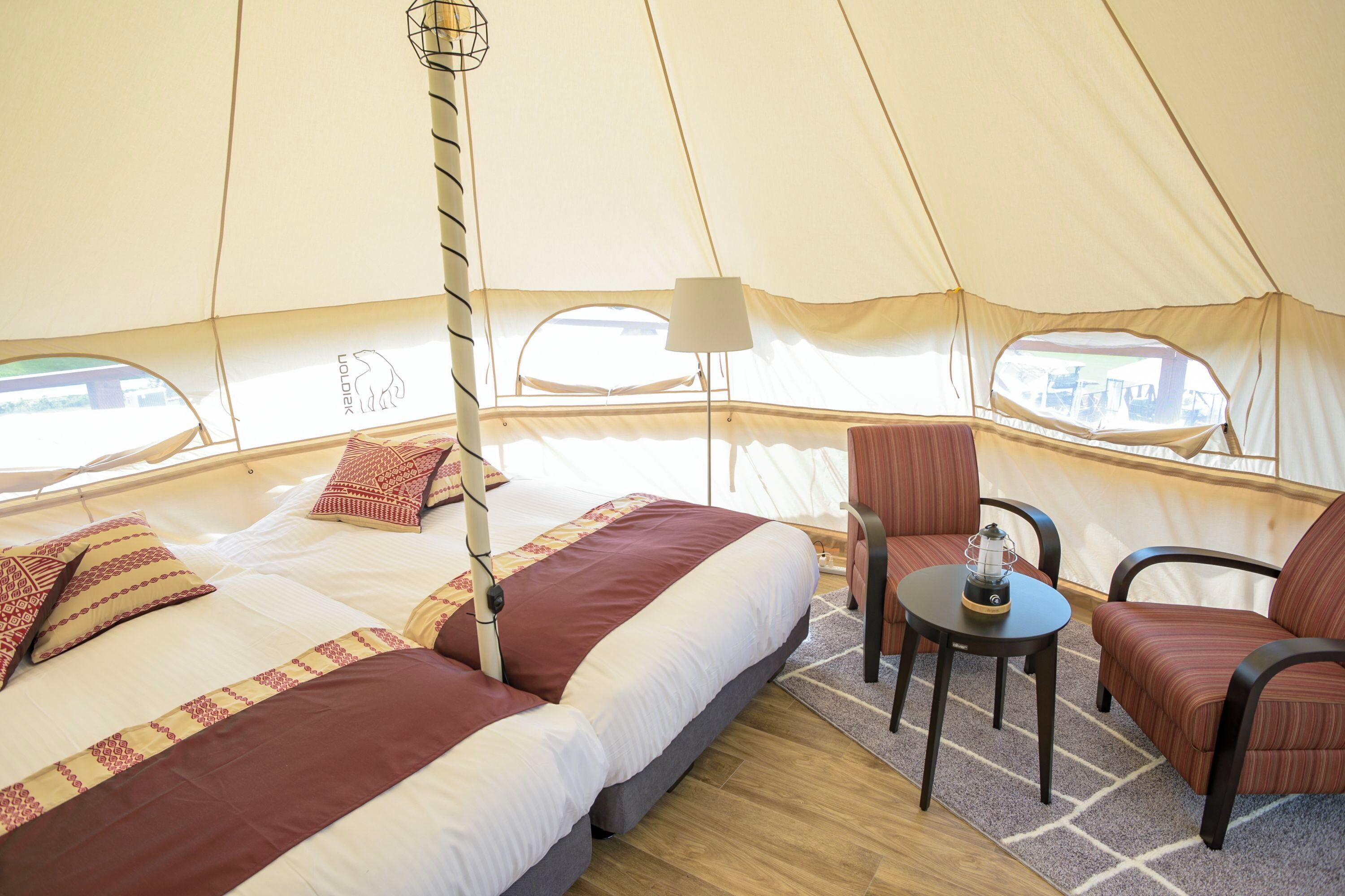 A tented accommodation with a bed, two chairs, a table, and a lamp.