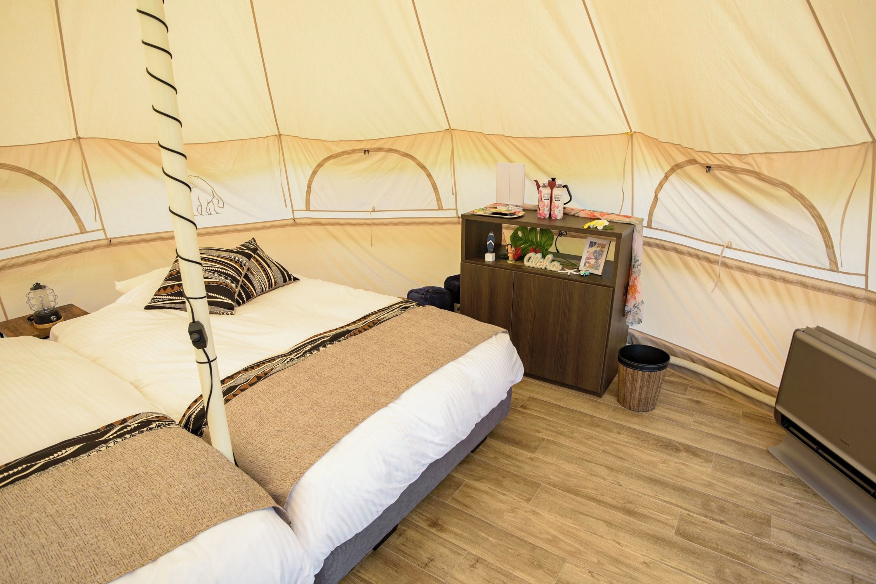 A tent with a bed, a wooden nightstand, a small table with a plant, and a trash bin.