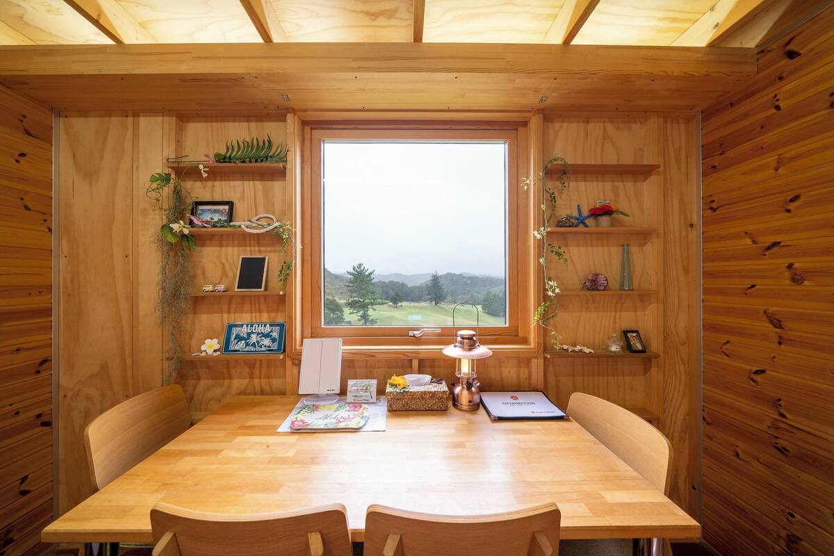 Een houten interieur met een eettafel, stoelen, een raam met uitzicht, plantenplanken en een lamp.