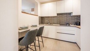 Une cuisine moderne avec des meubles blancs, un dosseret en carreaux noirs et une cuisinière encastrée. On y trouve deux tabourets de bar et un petit îlot central avec un saladier et des verres.