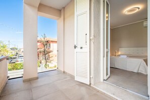 A balcony with a view of the street, a door leading to a bedroom with a bed and a nightstand, and a ceiling light fixture. - GuestHost - Mare Monti Apartment 7 (Dogana)