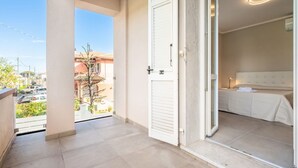 Un balcon donnant sur la rue, une porte menant à une chambre avec un lit et une table de chevet, et un luminaire au plafond.