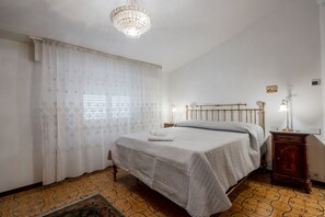 A bedroom with a bed, a chandelier, a wooden nightstand, and a patterned floor. - GuestHost - Mare Monti Apartment 8 (Dogana)