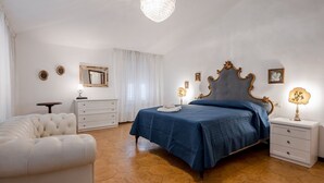Una camera da letto con un letto, una sedia bianca, un comodino, una cassettiera, un lampadario e uno specchio.