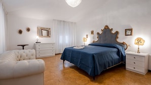 Een slaapkamer met een bed, een witte stoel, een nachtkastje, een dressoir, een kroonluchter en een spiegel.