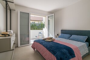 Un dormitorio con cama, mesita de noche, una silla y puerta corrediza de vidrio que da a un balcón exterior.