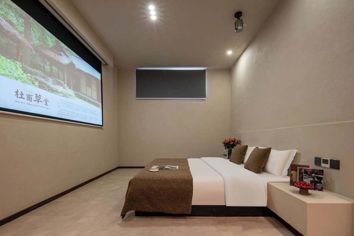Ein Hotelzimmer mit einem Bett, einer Projektionsleinwand, einem Fenster und einem Nachttisch mit einer Blumenvase.