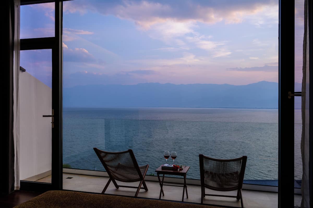 Ein Balkon mit zwei Korbstühlen und einem kleinen Tisch, auf dem zwei Weingläser und ein Buch stehen, mit Blick auf ein ruhiges Meer und Berge in der Ferne.