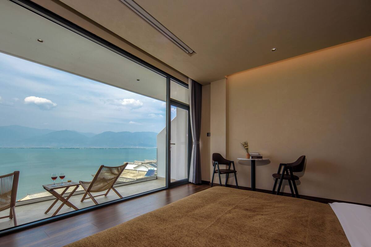 Ein modernes Hotelzimmer mit einer großen Schiebeglasdoor, die auf einen Balkon mit Blick auf Meer und Berge führt. Es gibt ein Bett, zwei Stühle und einen kleinen Tisch mit einem Blumenstrauß.