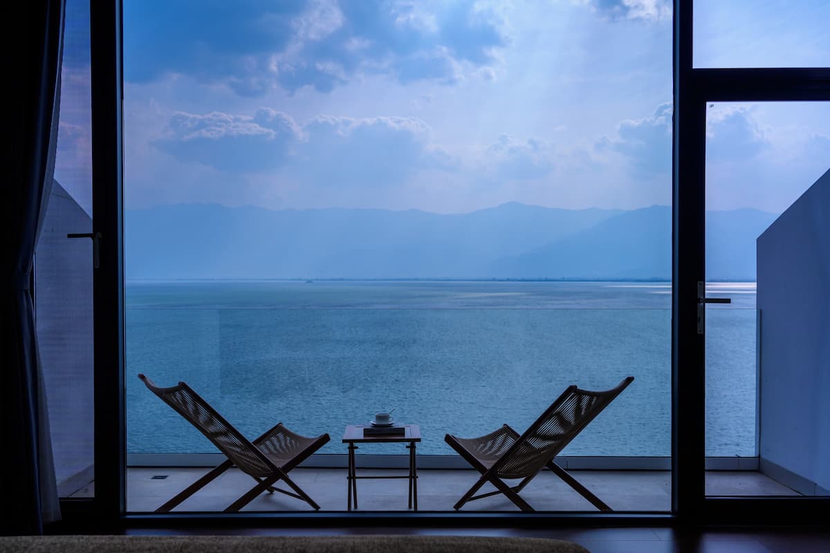 Ein Balkon mit zwei Korbstühlen und einem kleinen Tisch, mit Blick auf ein ruhiges Meer und ferne Berge.
