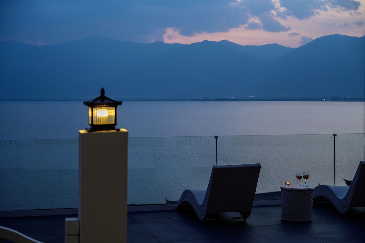 Ein Balkon mit Blick auf einen stillen See und Berge, eine Laterne sowie zwei Stühle mit einem kleinen Tisch und einer Kerze dazwischen.