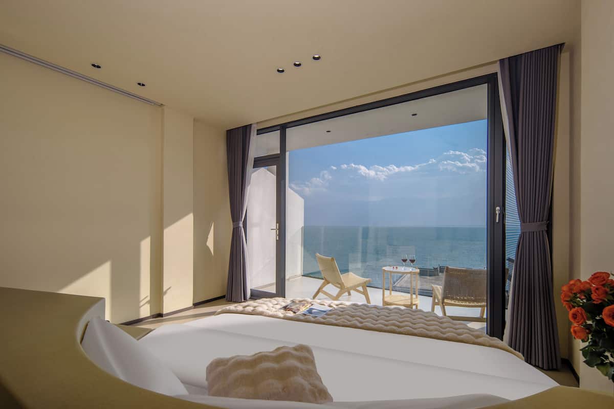 Ein Hotelzimmer mit einem großen Bett, Meerblick, einem kleinen Tisch mit einem Weinglas und einem Sessel am Fenster.