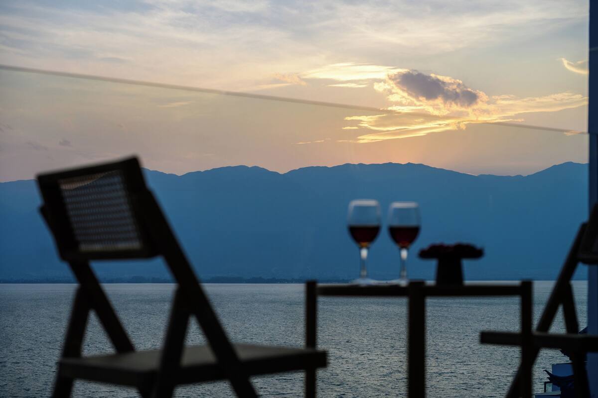 Ein Tisch mit zwei Weingläsern und einer kleinen Vase, mit Blick auf ein Gewässer und Berge im Hintergrund.