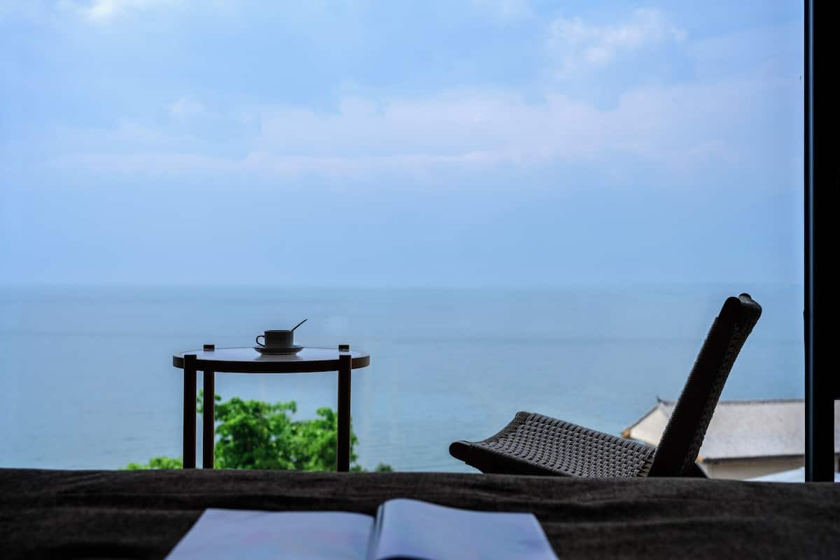 Blick vom Balkon auf das Meer, mit Tisch, Stuhl und offenem Buch auf einem Bett.