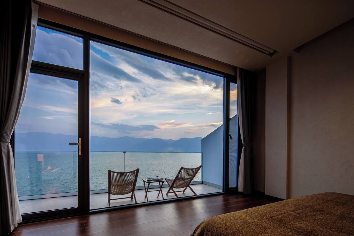 Ein Zimmer mit einer großen Schiebeglasdoor zum Balkon mit Blick auf Meer und Berge. Auf dem Balkon befinden sich zwei Korbstühle und ein kleiner Tisch.