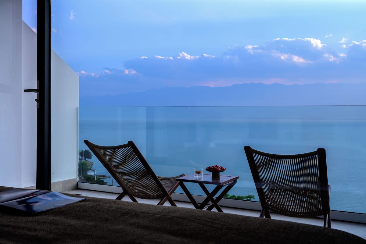 Ein Balkon mit zwei Korbstühlen und einem kleinen Tisch, mit Blick auf ein Gewässer und ferne Berge.