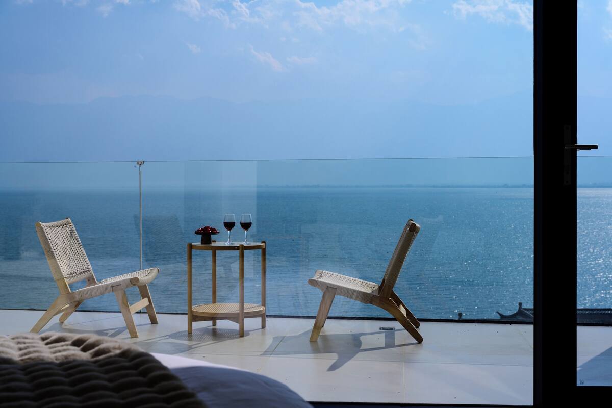 Ein Balkon mit zwei Korbstühlen und einem kleinen Tisch, auf dem zwei Gläser Rotwein stehen, mit Blick auf ein ruhiges Meer.