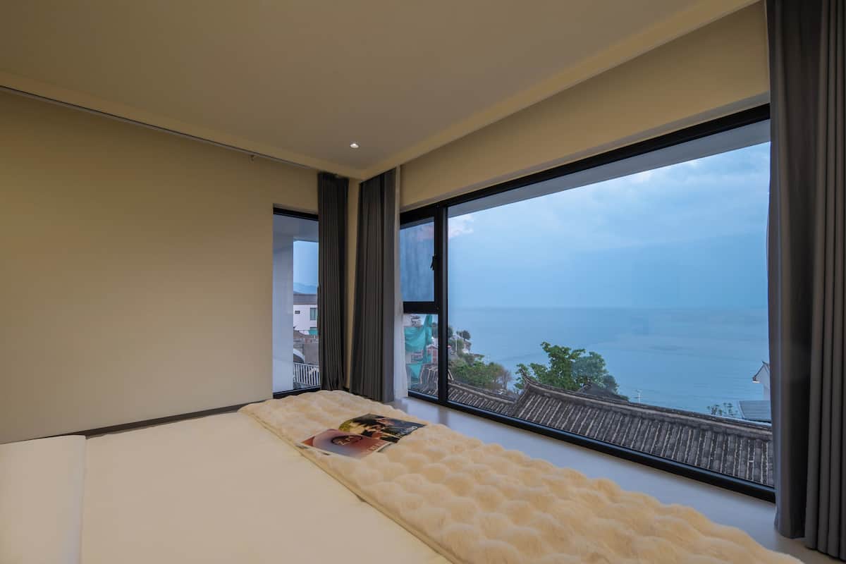 Ein Schlafzimmer mit großem Fenster mit Meerblick, einem Bett mit flauschiger weißer Bettdecke und einer Zeitschrift auf dem Bett.