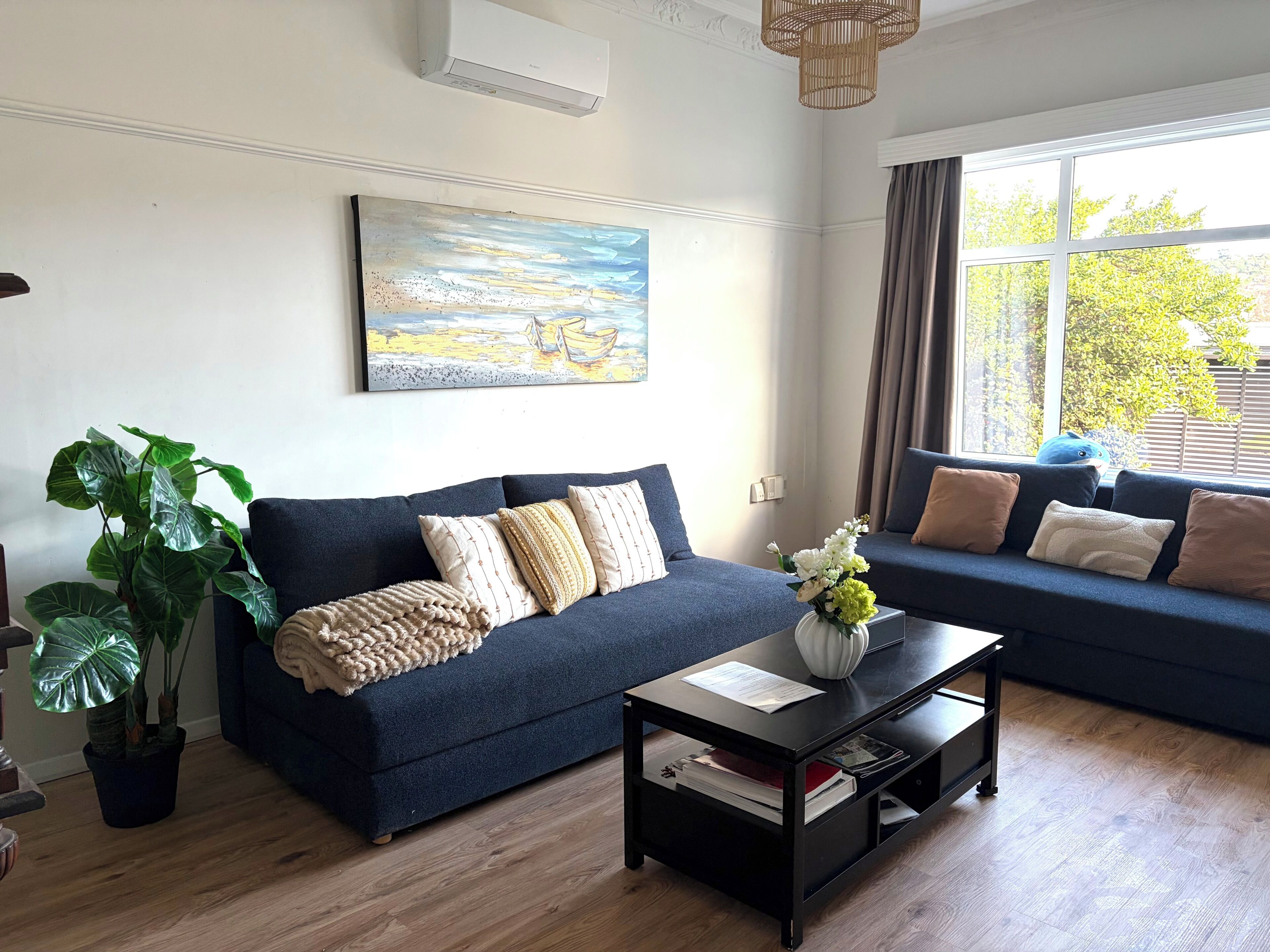 A living room with a dark blue sofa, a coffee table with a vase of flowers, and a large window with curtains.
