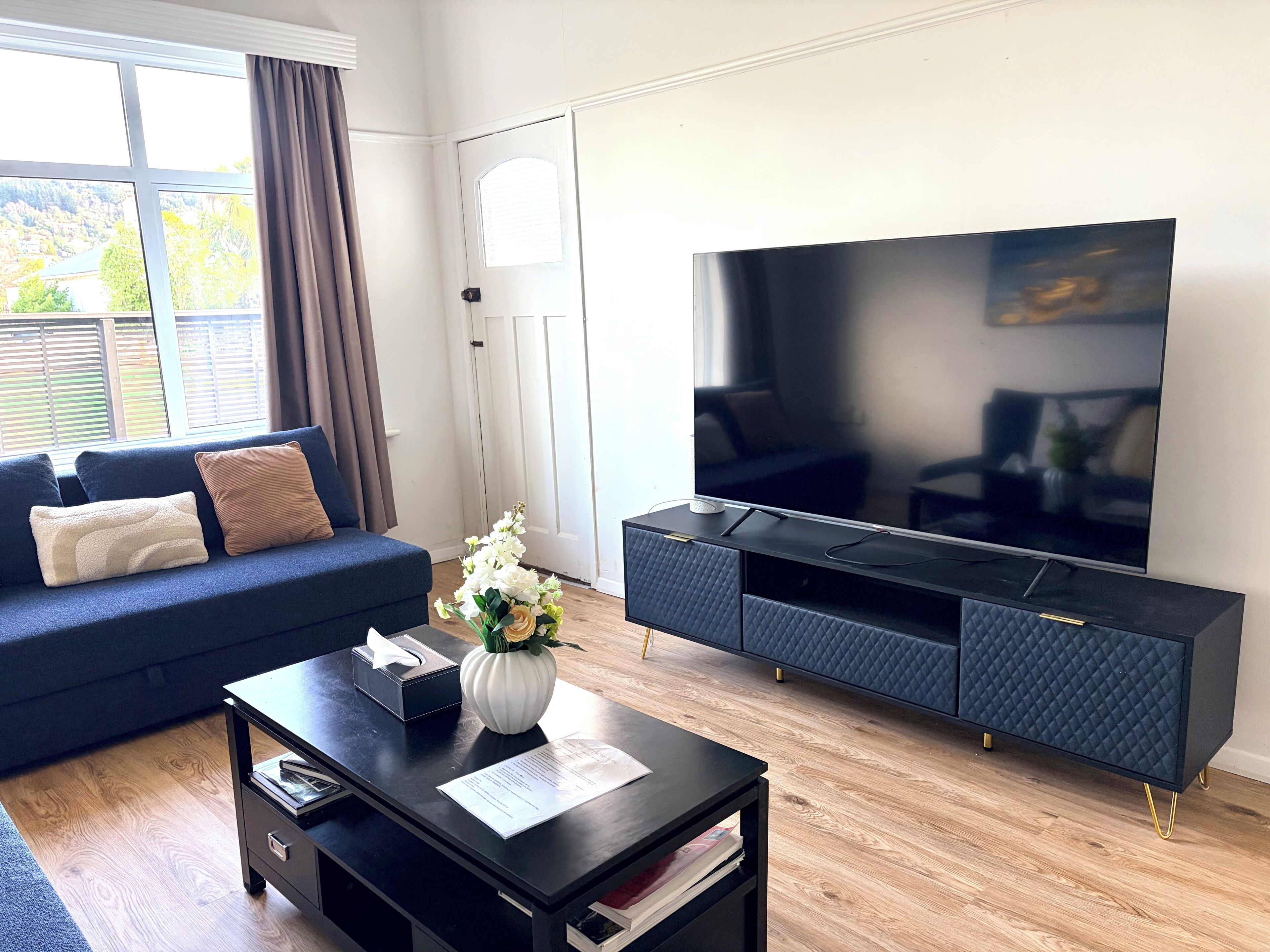 A modern living room with a large flat-screen TV, a dark blue sofa, a coffee table with a vase and papers, and a wooden floor.