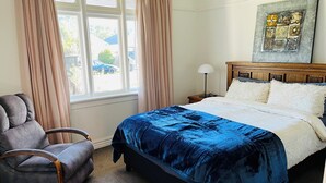 Ein Schlafzimmer mit Bett, Sessel, Nachttisch, Lampe und einem Fenster mit Vorhängen.