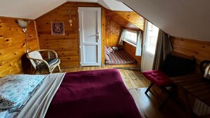 A cozy attic bedroom with a wooden interior, a bed with a red blanket, a chair, and a window with a view.