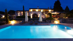 A swimming pool with lounge chairs and umbrellas, surrounded by stone walls and a lit-up building in the background.