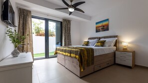 Un dormitorio con cama, mesita de noche, televisor, ventilador de techo y un cuadro en la pared.