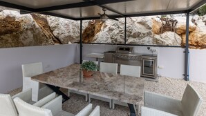 A covered outdoor dining area with a stone wall backdrop, a grill, and wicker chairs. - Villa Juma by Villas Guide (Blace)