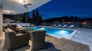Piscina con sillones de baño y mobiliario de mimbre, rodeada por un patio de piedra y vistas a árboles y edificios al anochecer.