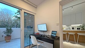 A modern office space with a desk, computer, and laptop. There is a sliding glass door leading to a balcony with a potted plant.