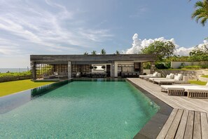 A modern pool area with a wooden deck, lounge chairs, and a bar area with a view of the ocean.