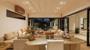 A modern living room with a large sofa set, a wooden coffee table, and a view of the outdoor garden at dusk.