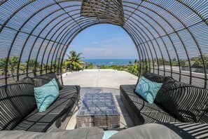 Un espacio de estar al aire libre con vistas al océano, que presenta una estructura metálica curva, dos sofás con cojines y una pequeña mesa.