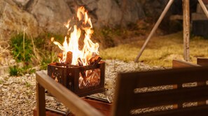 A metal fire pit with burning logs, surrounded by gravel and greenery. - Sokcho Supuro Caravan (Sokcho)