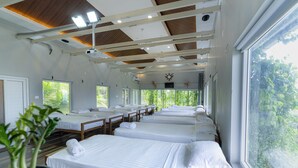 A row of neatly arranged beds with white linens, a ceiling with wooden beams, and large windows offering a view of greenery outside. - Floresta resorts (Idukki)