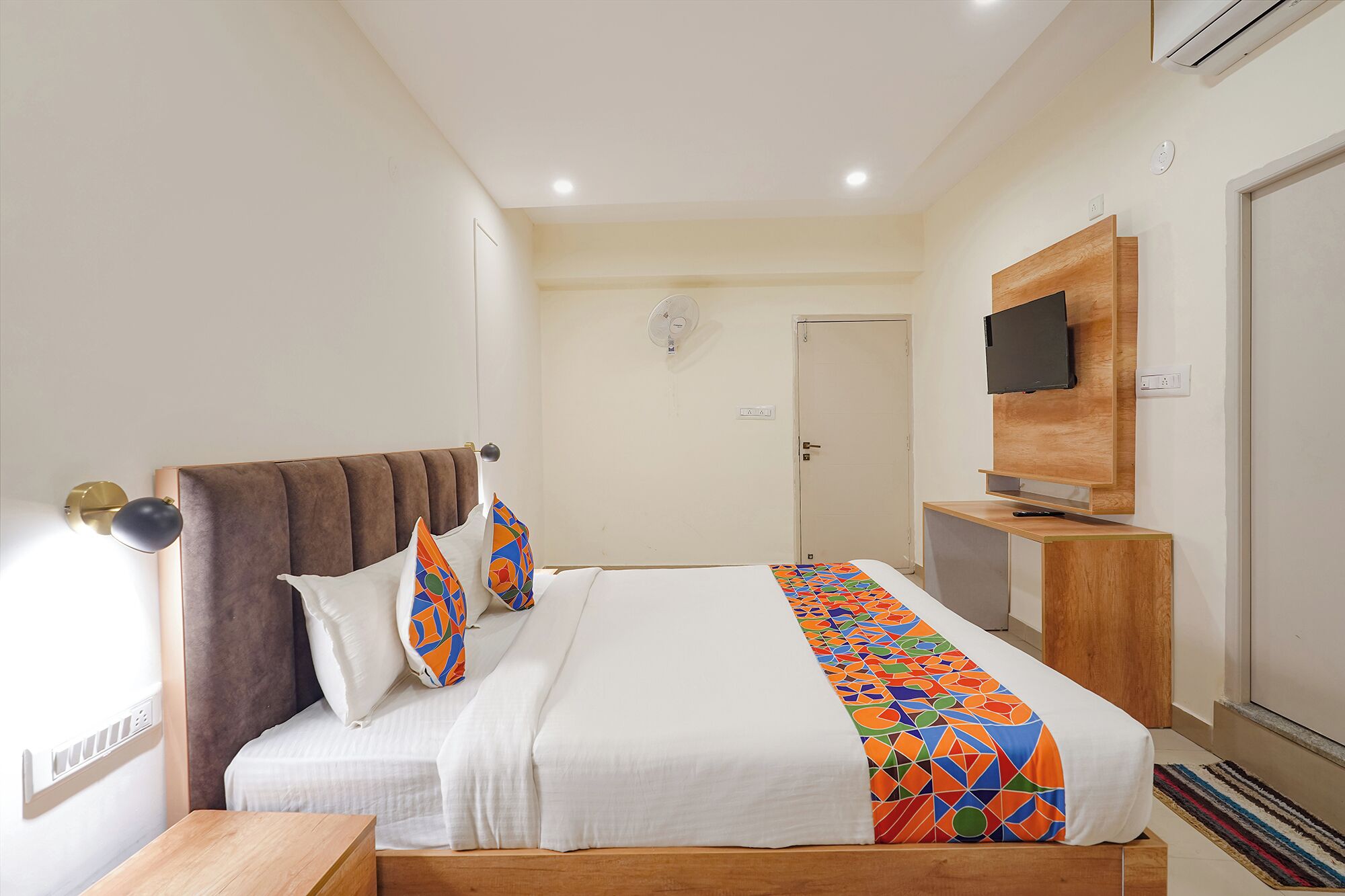 A modern hotel room with a large bed, a bedside table, a TV mounted on a wooden panel, and a door in the background.