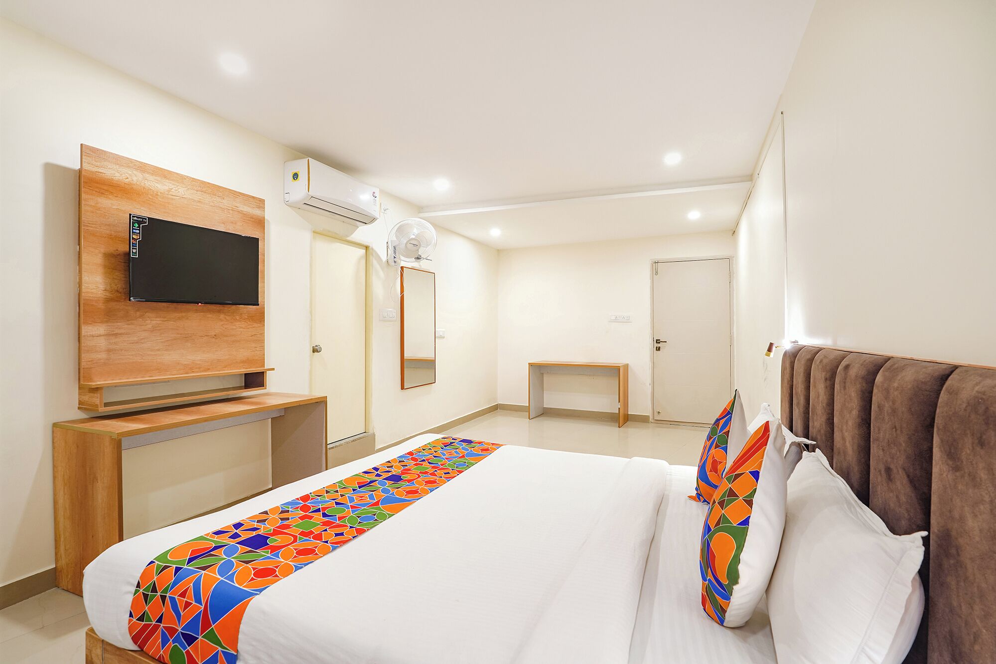 A modern hotel room with a bed, a TV mounted on a wooden panel, an air conditioning unit, a small desk, and a mirror.