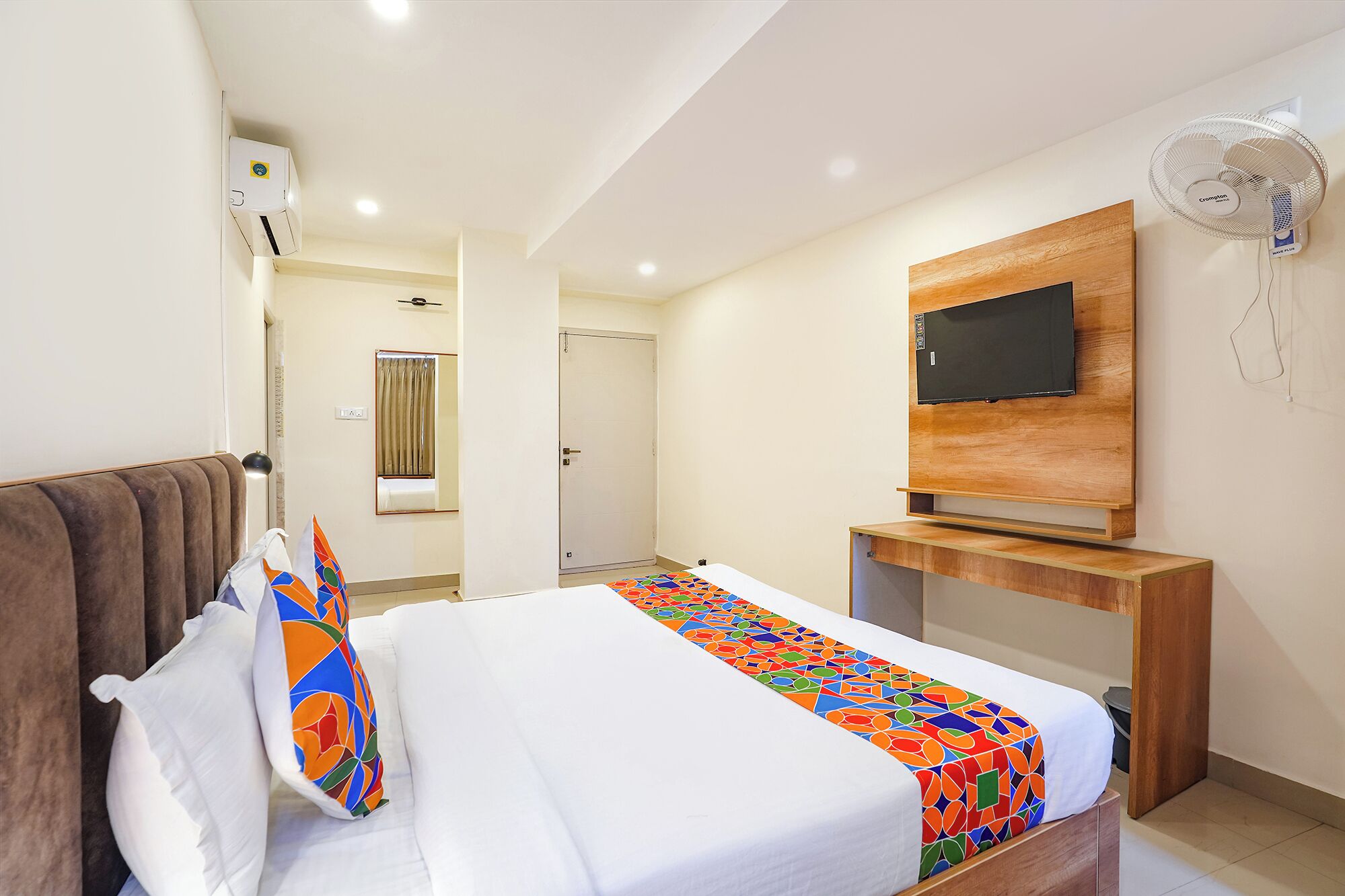 A hotel room with a bed, a TV mounted on a wooden panel, a ceiling fan, and a window with curtains.