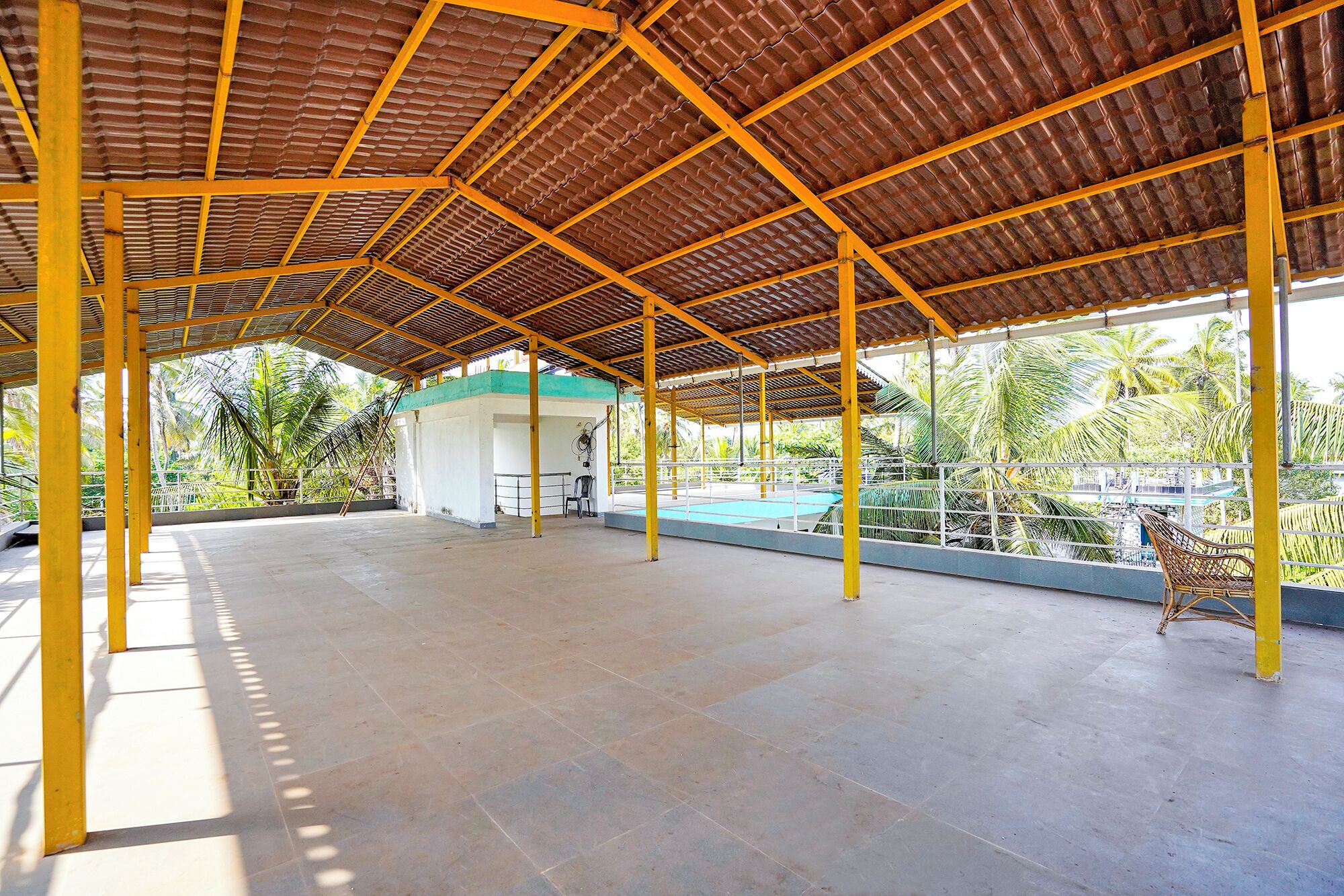 Uma área aberta coberta, com piso de azulejos, suportes estruturais amarelos e vista para palmeiras e um edifício ao fundo.