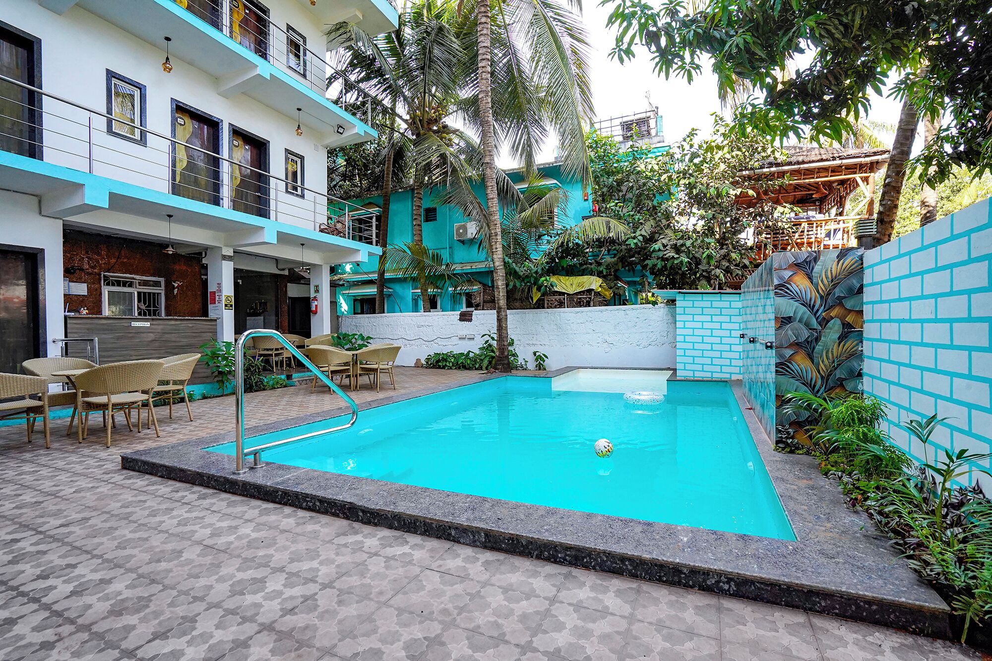 A swimming pool with a tiled deck, surrounded by a building with a balcony and a wall with a tropical leaf pattern.