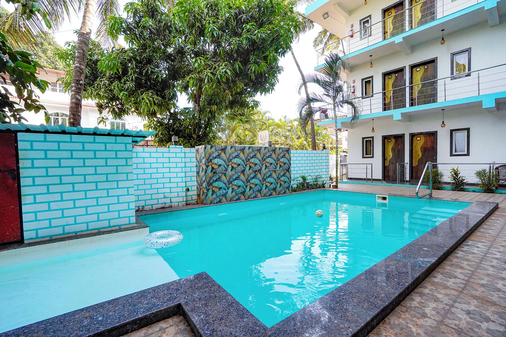 A swimming pool with a tiled border, a decorative wall, and a building with balconies and doors in the background.