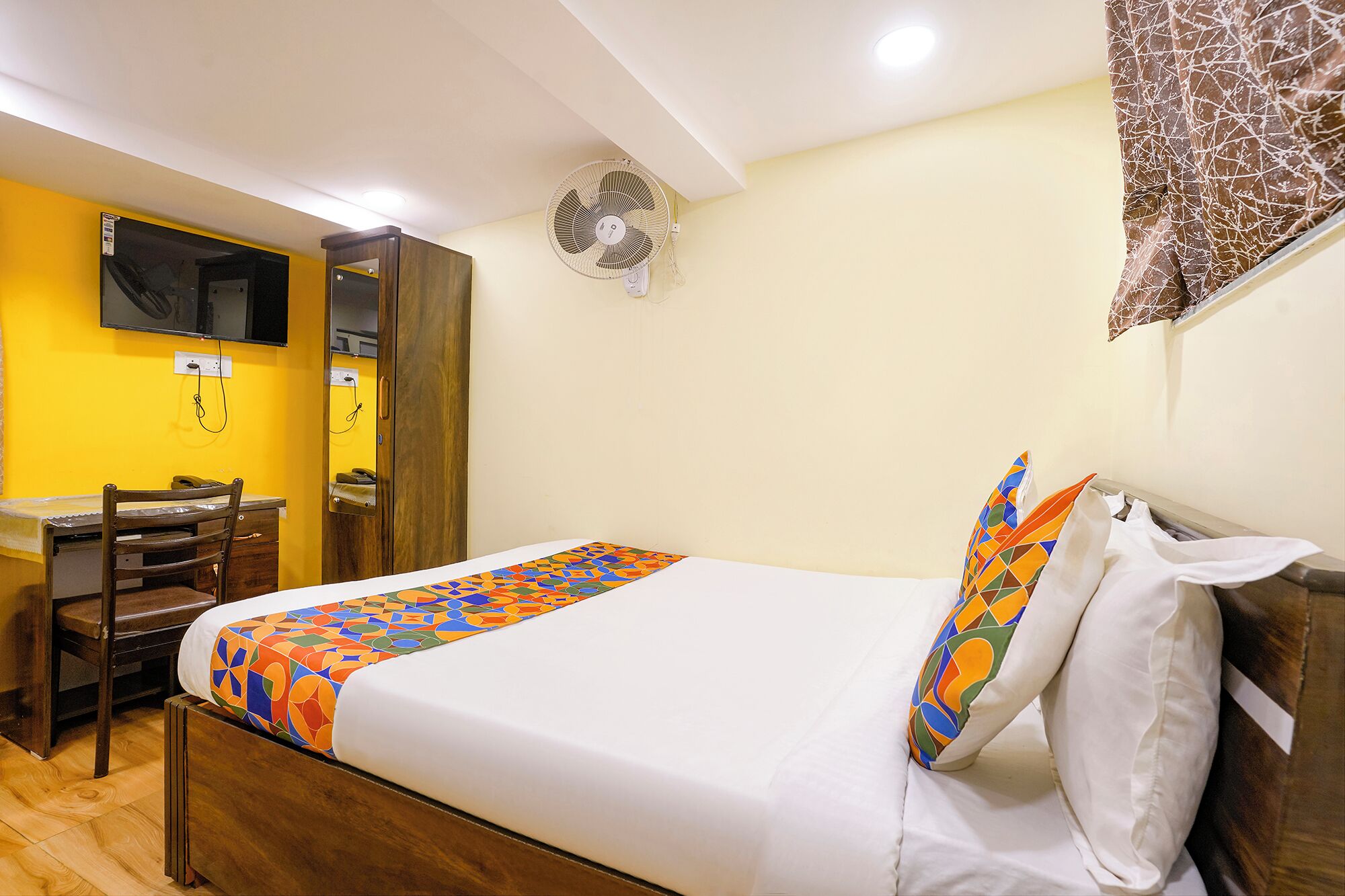 A hotel room with a bed, a desk, a chair, a TV mounted on the wall, and a ceiling fan.