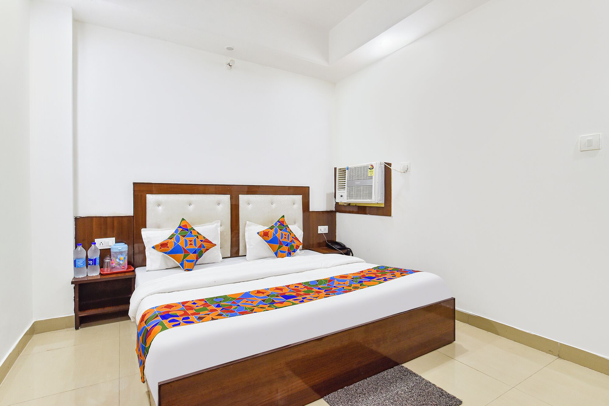 Een hotelkamer met een bed, nachtkastje, wandairconditioner en een klein tafeltje met waterflessen.