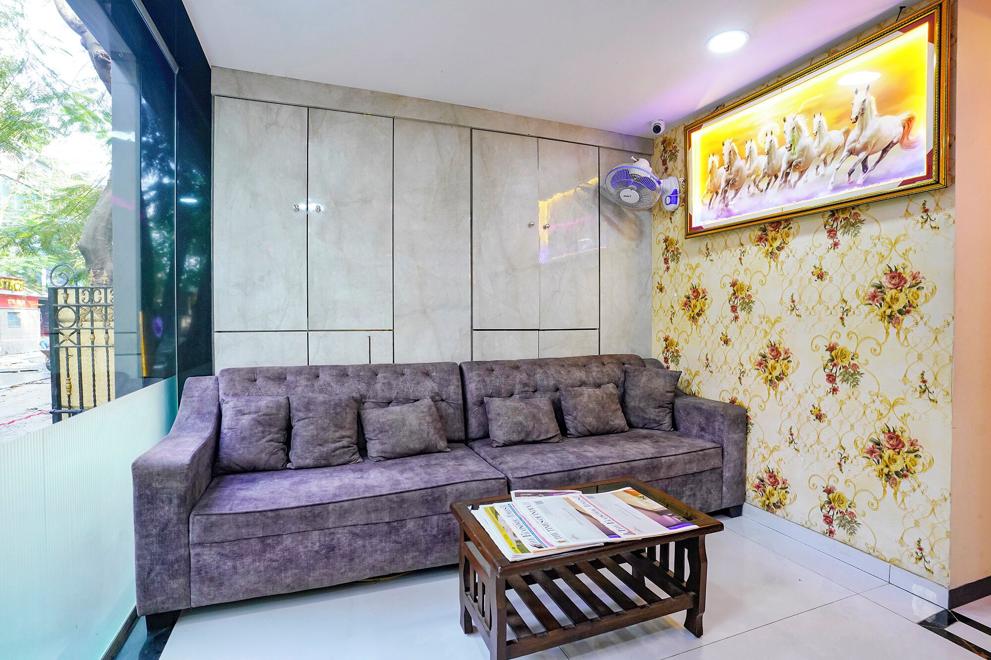 Una sala de estar moderna con un sofá gris, una mesa de centro de madera con revistas, un ventanal con vistas a árboles y un cuadro de caballos en la pared.